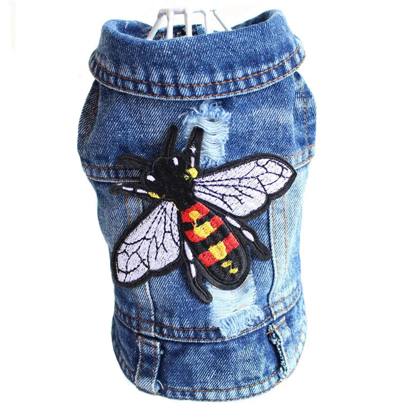 A blue denim pet jacket featuring a distressed, frayed hem and a large embroidered bee patch on the front, shown on a white hanger against a white background.